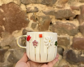 May include: A white ceramic mug with a gold handle and gold accents. The mug is decorated with colorful, raised floral designs. The flowers are red, orange, pink, white, and green. The mug is held in a hand.