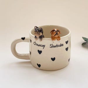 May include: Cream ceramic mug decorated with black hearts and the names "Chauncey" and "Shortcake." Two small dog figurines sit on the rim. The mug has a curved handle.
