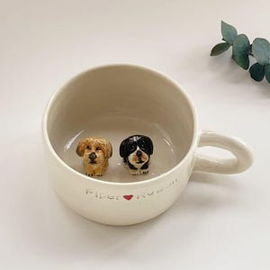 Könnte beinhalten: Cremefarbene Keramiktasse mit zwei kleinen Hundefiguren. Ein Hund ist hellbraun, der andere schwarz-weiß. Die Tasse hat einen Henkel und den Text "Piper ❤️ Rowan" in Schreibschrift.