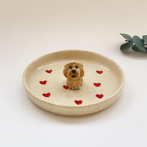May include: A round, speckled cream-colored ceramic trinket dish featuring a small, sculpted tan dog figurine in the center. The dish is decorated with several small, hand-painted red hearts scattered across the interior surface.
