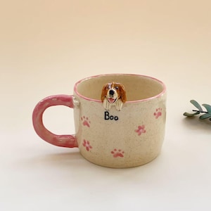 Puede incluir: Una taza de cer&aacute;mica color crema con asa y borde rosa, decorada con peque&ntilde;as huellas de patas rosas. Una figura esculpida de un perro marr&oacute;n y blanco asoma por el borde interior, sobre el texto negro "Boo" impreso en el exterior de la taza.