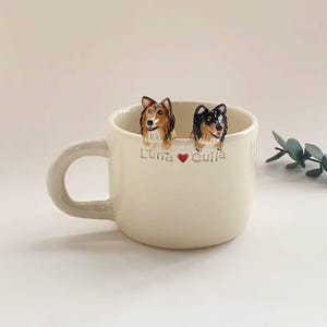 Op de afbeelding: Crèmekleurige keramische mok met twee hondenbeeldjes op de rand. Eén hond is bruin en bruin, de andere zwart en wit. De mok heeft een oor en de tekst "Luna ❤️ Quila" staat erop.