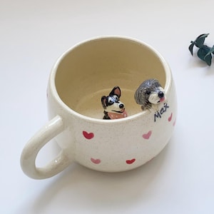 Puede incluir: Taza de cerámica color crema con asa, decorada con corazones rosas. Dos pequeñas figuras de perro están posadas en el borde. Una es negra y blanca con una bufanda rosa, la otra es gris. El nombre "Max" está escrito en el lateral.