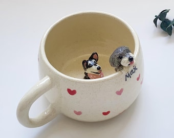 Puede incluir: Taza de cerámica color crema con asa, decorada con corazones rosas. Dos pequeñas figuras de perro están posadas en el borde. Una es negra y blanca con una bufanda rosa, la otra es gris. El nombre "Max" está escrito en el lateral.