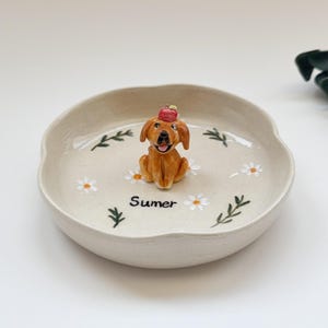 May include: A beige ceramic dish with a scalloped edge, decorated with daisies and green leaves. A small, brown dog figurine wearing a red hat sits in the centre. The word "Sumer" is written on the dish.