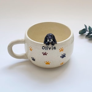 May include: Cream-colored ceramic mug with a black and white dog figurine perched on the rim. The mug is decorated with paw prints in orange, brown, and blue, and the name "Olivia" is written on the side.