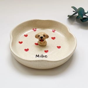 May include: A beige ceramic pet dish featuring a golden retriever figurine. The dish is decorated with red hearts and the name "Millie". The dish is ideal for a small dog or cat.
