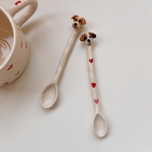 Könnte beinhalten: Zwei Keramik-Löffel mit Hunde-Kopf-Details. Ein Löffel hat rote und rosa Herz-Akzente. Eine passende Tasse mit roten Herz-Akzenten ist ebenfalls sichtbar. Die Löffel und die Tasse sind cremefarben.