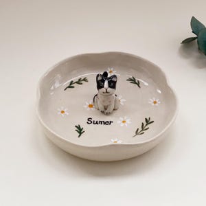 Op de afbeelding: Een lichtbeige keramische schaal met een zwart-witte kattenfiguur, witte madeliefjes en groene takjes. Het woord "Sumer" staat op de schaal. De schaal heeft een katten thema.
