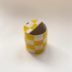 May include: A cylindrical ceramic container featuring a hand-painted yellow and white checkerboard pattern. The vessel includes a partial lid covering one side of the top opening, which features a small circular cutout for a spoon or utensil.