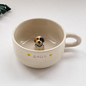 Könnte beinhalten: Eine weiße Keramik-Tasse mit schwarzem Henkel und dem Text "But First, Coffee" in fetter, schwarzer Schrift. Die Tasse steht vor einem verschwommenen Hintergrund, der eine gemütliche Umgebung suggeriert.