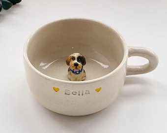 Puede incluir: Una taza de cerámica de color crema con una pequeña figura tridimensional de un perro con un collar de lunares azules en el fondo. El exterior de la taza tiene la inscripción Bella y dos pequeños corazones amarillos.