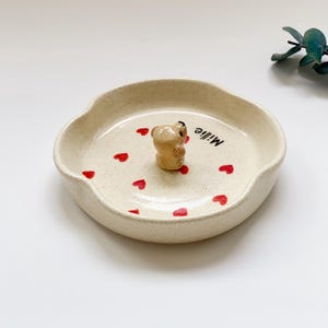 May include: A beige ceramic dish with a scalloped edge, featuring a small bear figurine in the centre. Red heart shapes are scattered around the bear. The word "Millie" is written in black script on the dish.