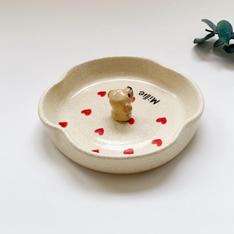 May include: A beige ceramic dish with a scalloped edge, featuring a small bear figurine in the centre. Red heart shapes are scattered around the bear. The word "Millie" is written in black script on the dish.
