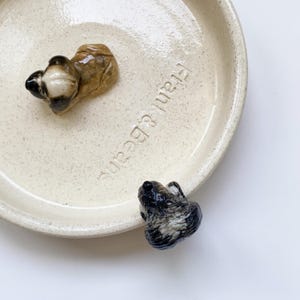 May include: A cream-colored ceramic dish with the text "Frank & Beans" and two small, decorative animal figurines. One figurine is brown and tan, the other is black and white. The dish has a speckled texture.
