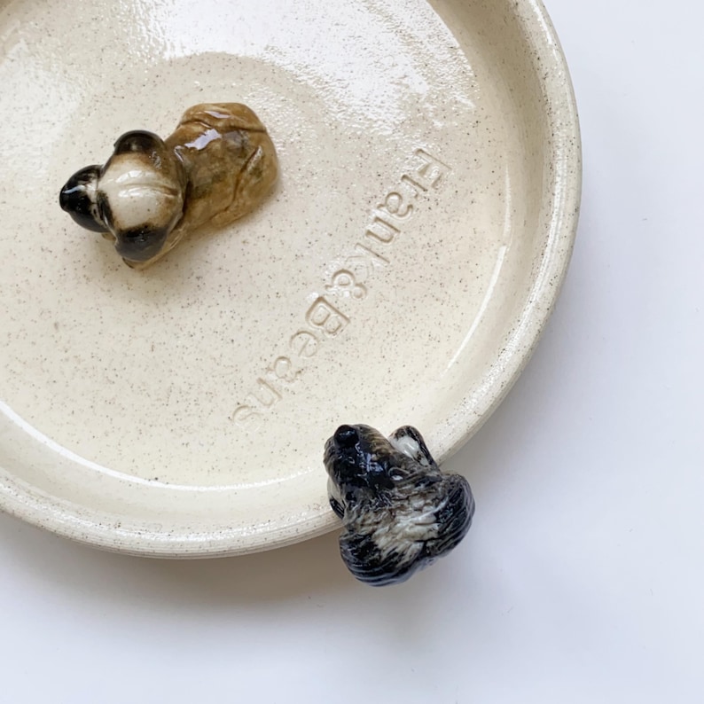 May include: A cream-colored ceramic dish with the text "Frank & Beans" and two small, decorative animal figurines. One figurine is brown and tan, the other is black and white. The dish has a speckled texture.