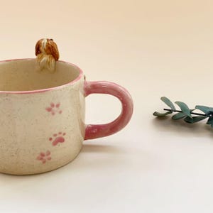 Puede incluir: Una taza de cer&aacute;mica con acabado crema moteado, con un borde rosa y un asa rosa a juego. El exterior est&aacute; decorado con peque&ntilde;as huellas de patas rosas, y una peque&ntilde;a figura esculpida de un perro marr&oacute;n y blanco est&aacute; unida al borde interior.