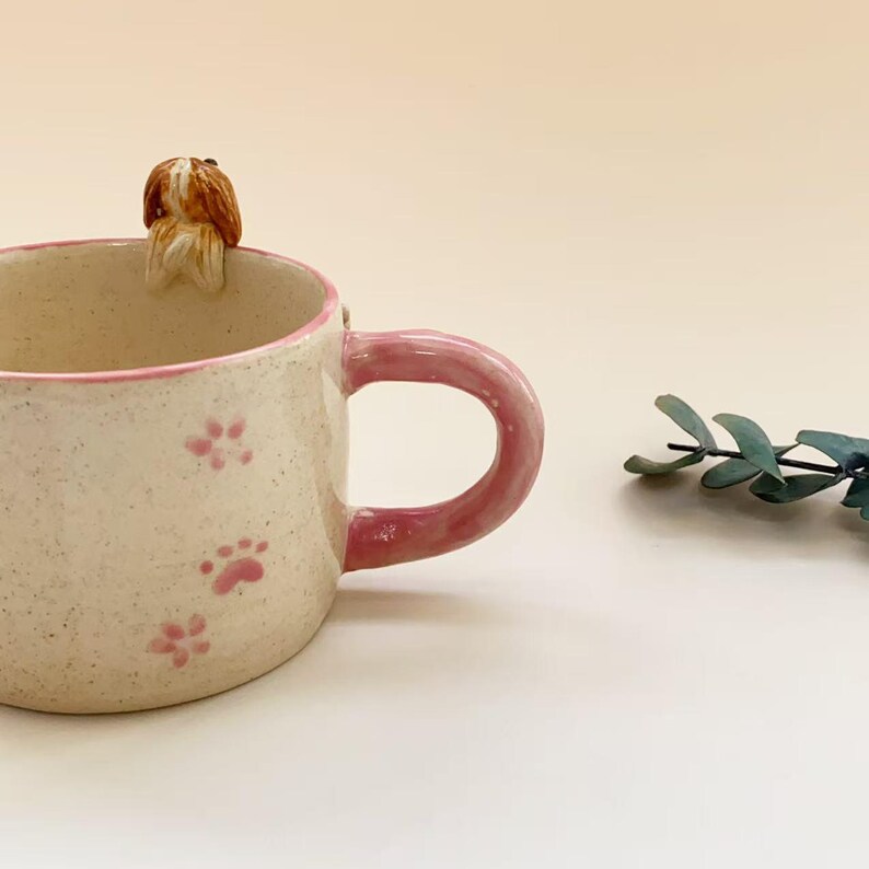 Puede incluir: Una taza de cer&aacute;mica con acabado crema moteado, con un borde rosa y un asa rosa a juego. El exterior est&aacute; decorado con peque&ntilde;as huellas de patas rosas, y una peque&ntilde;a figura esculpida de un perro marr&oacute;n y blanco est&aacute; unida al borde interior.
