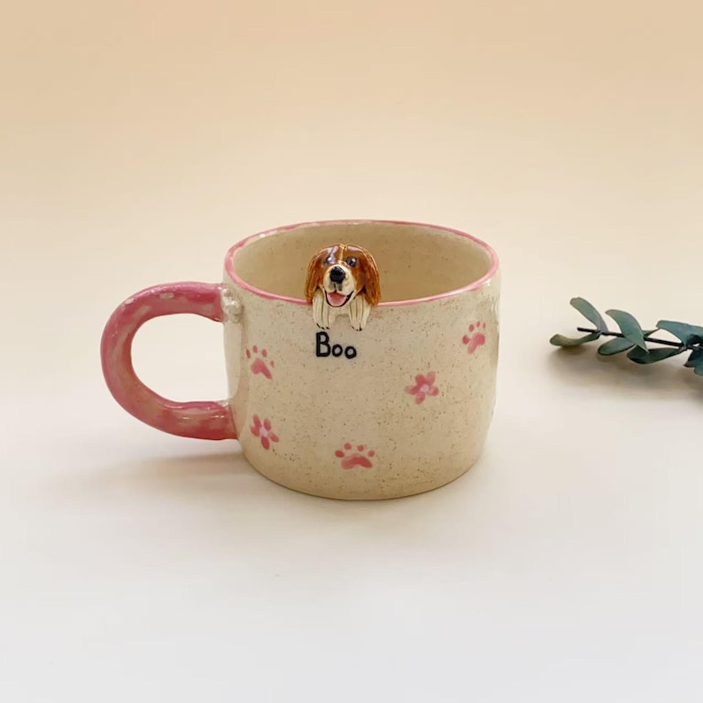 Puede incluir: Una taza de cer&aacute;mica color crema con asa y borde rosa, decorada con peque&ntilde;as huellas de patas rosas. Una figura esculpida de un perro marr&oacute;n y blanco asoma por el borde interior, sobre el texto negro "Boo" impreso en el exterior de la taza.
