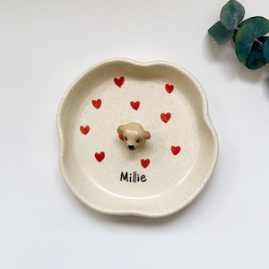 May include: A beige ceramic dish with a wavy edge, decorated with red hearts and a small, tan dog figurine in the centre. The name "Millie" is written in black script at the bottom. A charming, decorative piece.