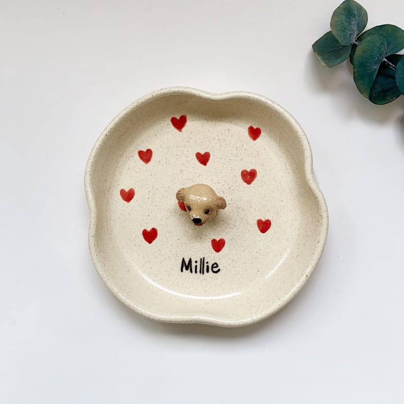 May include: A beige ceramic dish with a wavy edge, decorated with red hearts and a small, tan dog figurine in the centre. The name "Millie" is written in black script at the bottom. A charming, decorative piece.