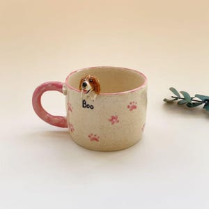 Puede incluir: Una taza de cer&aacute;mica color crema con acabado moteado, asa rosa y patrones de huellas de patas rosas. Una peque&ntilde;a figura tridimensional de un perro marr&oacute;n y blanco est&aacute; fijada al borde, con la palabra "Boo" impresa en negro debajo.