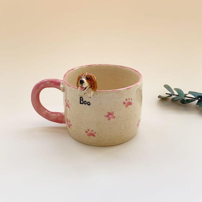 Puede incluir: Una taza de cer&aacute;mica color crema con acabado moteado, asa rosa y patrones de huellas de patas rosas. Una peque&ntilde;a figura tridimensional de un perro marr&oacute;n y blanco est&aacute; fijada al borde, con la palabra "Boo" impresa en negro debajo.