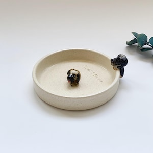 May include: A beige ceramic dog bowl with two small dog figurines. One dog is in the center of the bowl, and the other is perched on the edge. The bowl has the text "Frank & Bear" on the side.