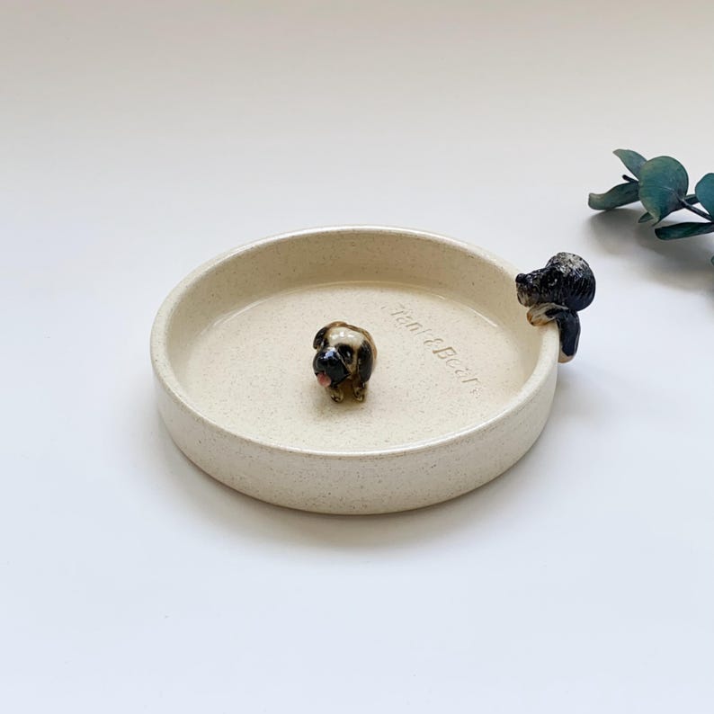 May include: A beige ceramic dog bowl with two small dog figurines. One dog is in the center of the bowl, and the other is perched on the edge. The bowl has the text "Frank & Bear" on the side.