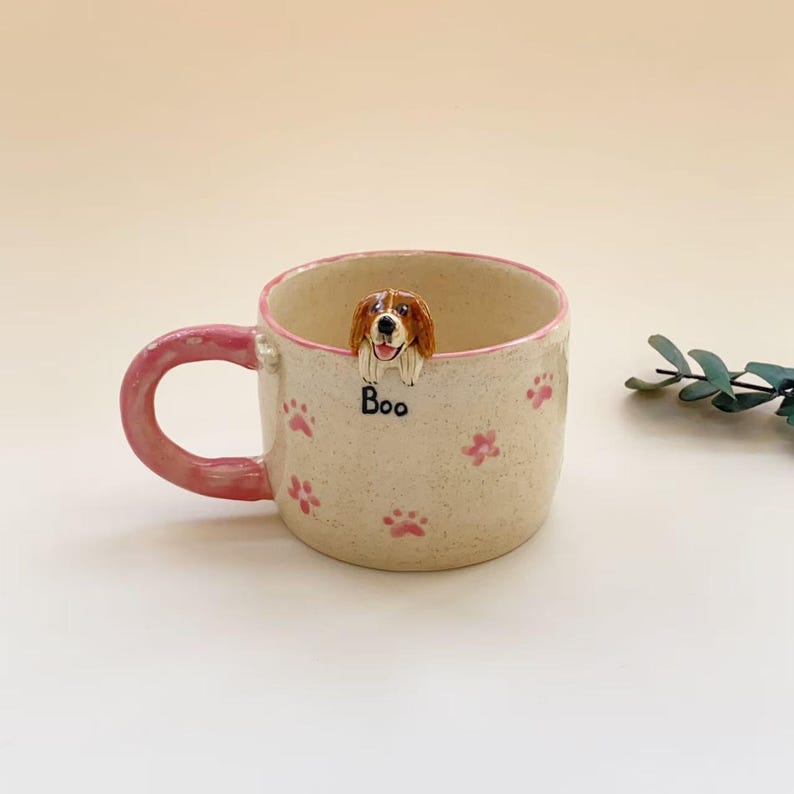 Puede incluir: Una taza de cer&aacute;mica con acabado crema moteado, con asa y borde de color rosa. El exterior est&aacute; decorado con peque&ntilde;as huellas de patas rosas y una cabeza de perro esculpida en marr&oacute;n y blanco que asoma por el borde, con la palabra Boo en negro.
