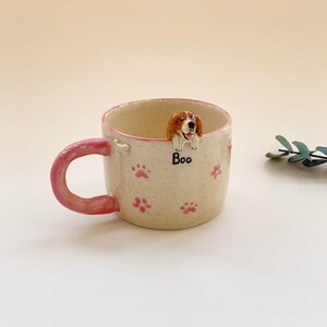 Puede incluir: Una taza de cer&aacute;mica color crema con motas, asa y borde rosa. El exterior presenta huellas de patas rosas y la palabra "Boo" escrita en negro. Una figura tridimensional de un perro marr&oacute;n y blanco est&aacute; fijada en el borde interior.
