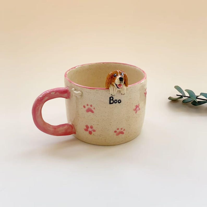 Puede incluir: Una taza de cer&aacute;mica color crema con motas, asa y borde rosa. El exterior presenta huellas de patas rosas y la palabra "Boo" escrita en negro. Una figura tridimensional de un perro marr&oacute;n y blanco est&aacute; fijada en el borde interior.
