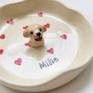 May include: A cream ceramic pet bowl featuring a dog figurine and red heart decorations. The bowl has the name "Millie" written in blue script. The bowl is ideal for a small pet.