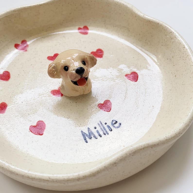 May include: A cream ceramic pet bowl featuring a dog figurine and red heart decorations. The bowl has the name "Millie" written in blue script. The bowl is ideal for a small pet.