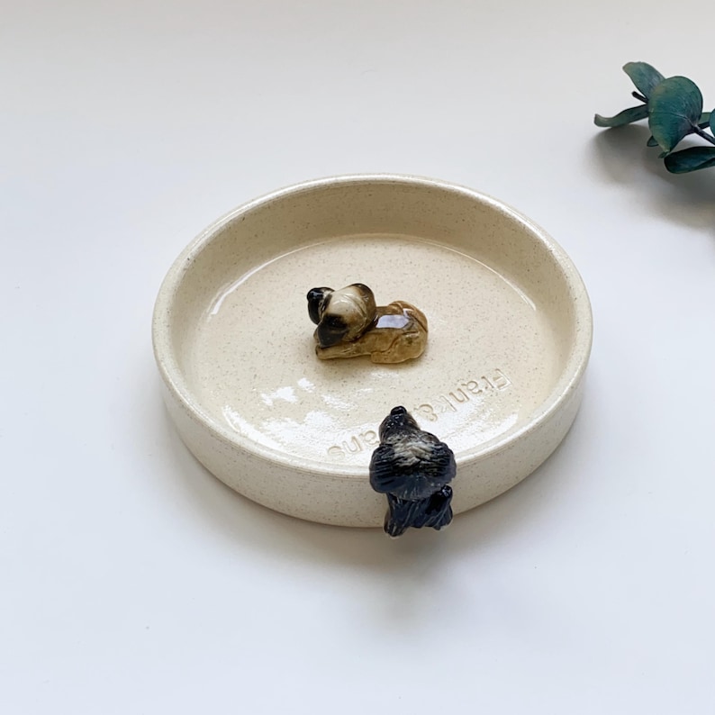 May include: A beige ceramic dish with two small animal figurines. One figurine is a brown and tan dog, and the other is a black bird. The dish has the text "Frank & Beans" on the side.