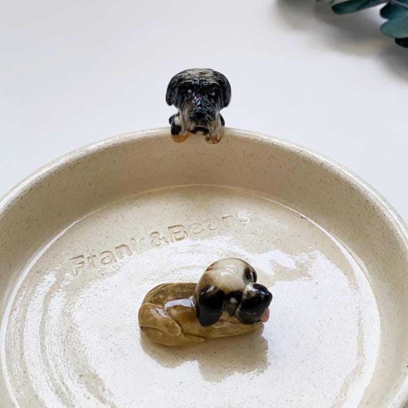 May include: A beige ceramic dog bowl with the text "Frank & Beans" embossed on the side. The bowl features two small ceramic dog figurines, one perched on the rim and the other lying down inside the bowl.