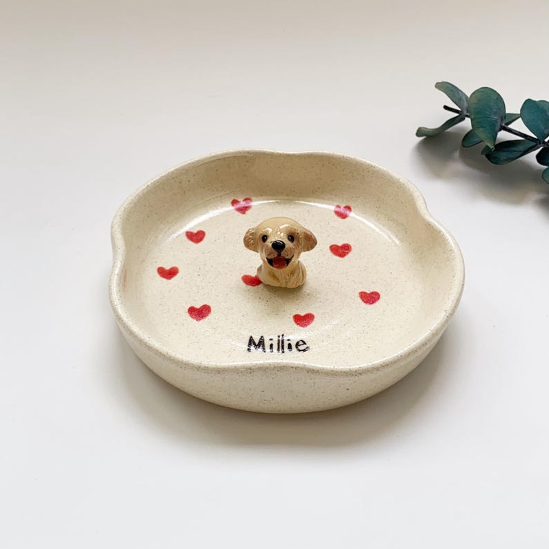 May include: A beige ceramic pet dish featuring a golden retriever figurine. The dish is decorated with red hearts and the name "Millie". The dish is ideal for a small dog or cat.