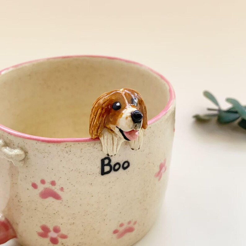 Puede incluir: Una taza de cer&aacute;mica moteada con borde rosa, que presenta una figura esculpida de un perro marr&oacute;n y blanco asom&aacute;ndose por el borde. La taza est&aacute; decorada con peque&ntilde;as huellas de patas rosas y la palabra Boo en letras negras bajo el perro.