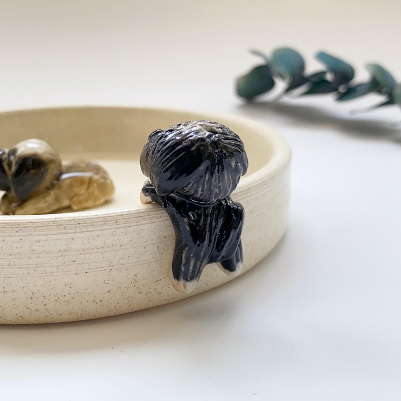 May include: A ceramic bowl with two small dog figurines. One dog is brown and tan, lying down. The other is black and white, appearing to hang over the edge of the bowl. The bowl is a light beige color.