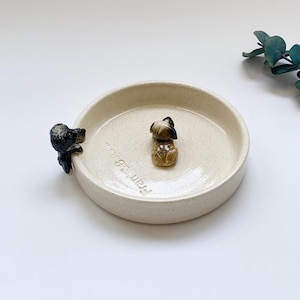 May include: A beige ceramic dish with two small dog figurines. One dog is perched on the edge, and the other is in the center. The dish has the text "Frank & Bear" on the side.