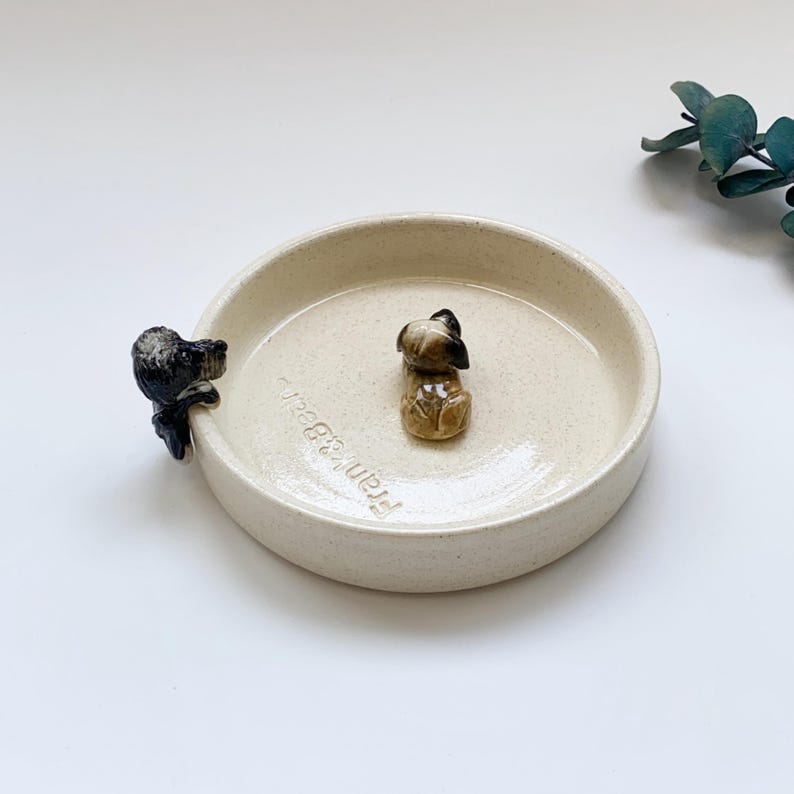 May include: A beige ceramic dish with two small dog figurines. One dog is perched on the edge, and the other is in the center. The dish has the text "Frank & Bear" on the side.