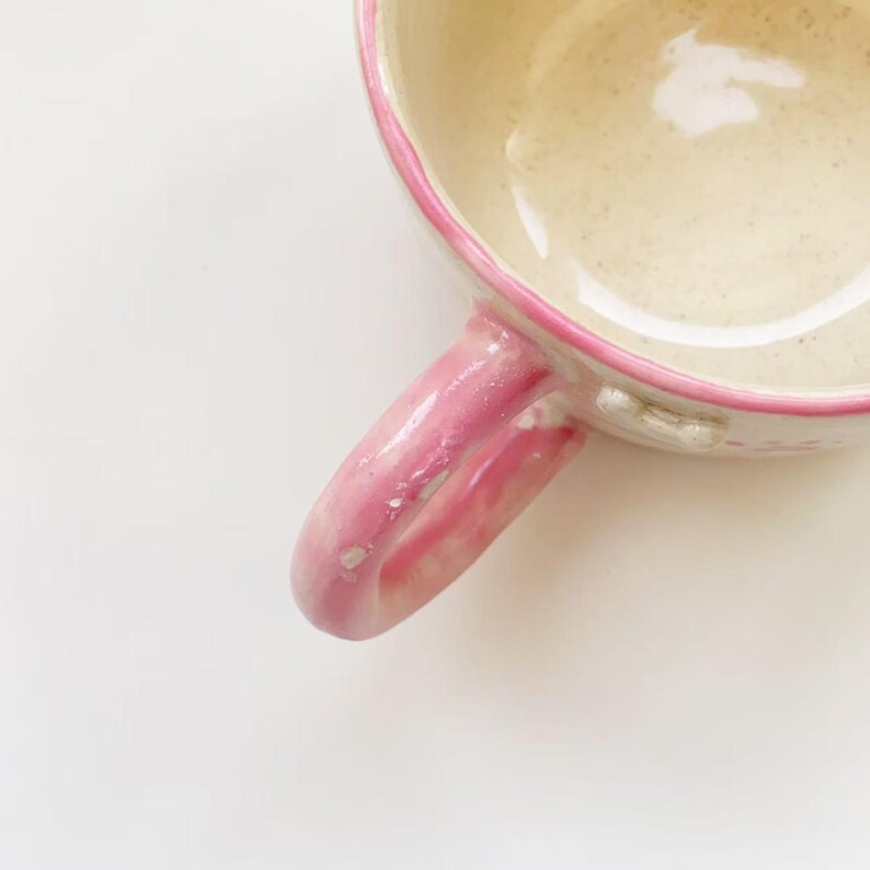 Puede incluir: Una taza de cer&aacute;mica con un interior de color crema brillante con motas sutiles y un borde esmaltado en rosa vibrante. El asa tambi&eacute;n est&aacute; acabada con un esmalte rosa brillante a juego, mostrando una forma redondeada y una textura artesanal.