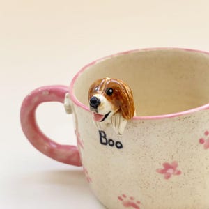 Puede incluir: Una taza de cer&aacute;mica con acabado crema moteado, borde y asa de color rosa. El exterior est&aacute; decorado con peque&ntilde;as flores y huellas de patas rosas. Una figura tridimensional de un perro marr&oacute;n y blanco est&aacute; fijada al borde, con la palabra Boo.