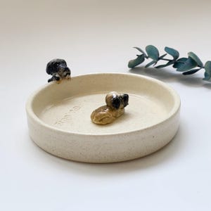 May include: A cream-colored ceramic pet bowl with two small dog figurines. One dog is perched on the edge, and the other is lying down inside the bowl. The bowl has a speckled texture and a slightly raised rim.
