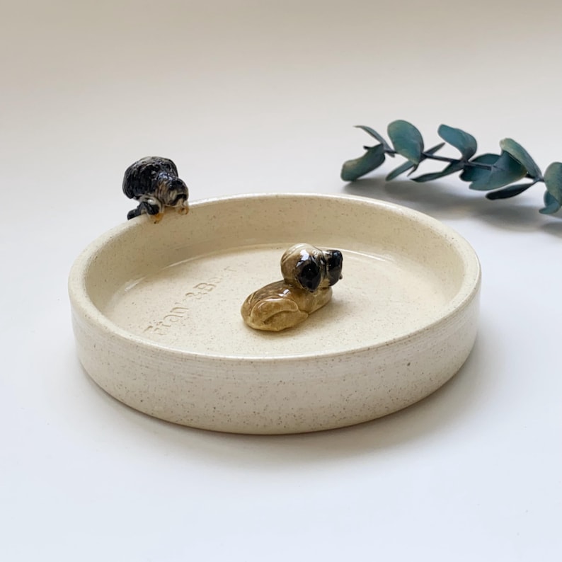 May include: A cream-colored ceramic pet bowl with two small dog figurines. One dog is perched on the edge, and the other is lying down inside the bowl. The bowl has a speckled texture and a slightly raised rim.