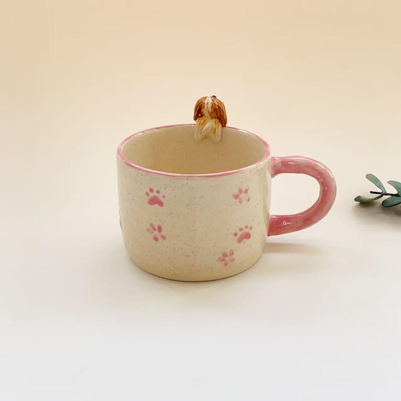 Puede incluir: Una taza de cer&aacute;mica con acabado crema moteado, borde rosa y asa rosa a juego. El exterior est&aacute; decorado con peque&ntilde;as huellas de patas rosas, y una peque&ntilde;a figura tridimensional de un perro marr&oacute;n y blanco se asienta en el borde interior.