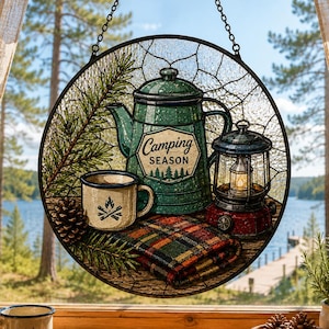 Könnte beinhalten: Ein runder Buntglas-Sonnenfänger mit einer türkisfarbenen Kaffeekanne mit der Aufschrift Camping Season, einer cremefarbenen Tasse mit Lagerfeuer-Symbol, einer roten Laterne, einer karierten Decke und Tannenzweigen.