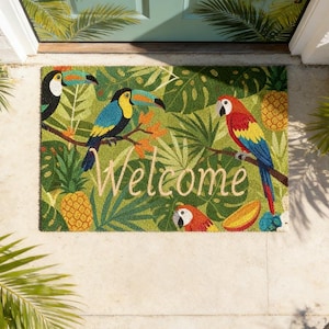 Op de afbeelding: Een rechthoekige deurmat met een tropisch thema. De mat heeft een groene achtergrond met illustraties van toekans, papegaaien, ananassen en tropische bladeren. Het woord "Welcome" staat in lichtbruin in het midden.