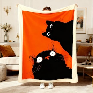 May include: A large, rectangular blanket featuring a vibrant orange background with two black cat illustrations. The cats have large, wide eyes. The blanket has a cream-colored border.