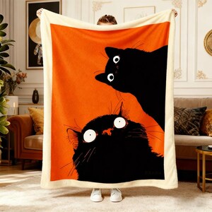 May include: A large orange blanket featuring two black cartoon cats with wide, surprised eyes. The blanket has a cream-colored border and a soft, plush texture, perfect for snuggling. The cats are set against a vibrant orange background.